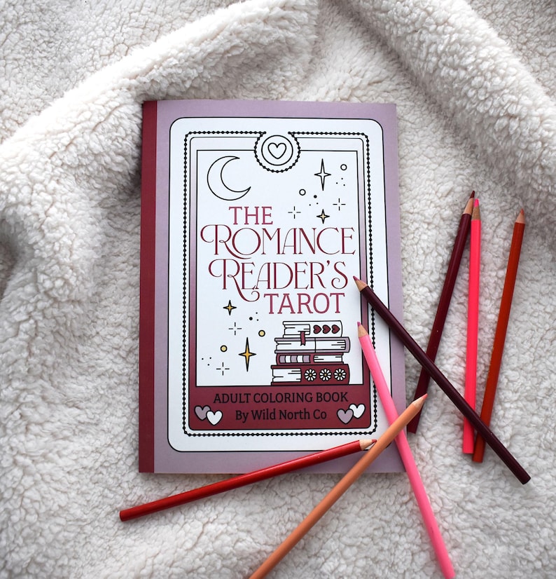 May include: A purple and red coloring book with the title "The Romance Reader's Tarot" and the text "Adult Coloring Book by Wild North Co". The book is surrounded by colored pencils on a white fluffy surface.