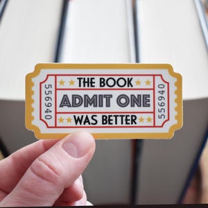 May include: A yellow and white sticker shaped like a movie ticket with the text "The Book Was Better" and "Admit One" in gray text. The ticket number is 556940.