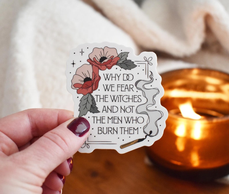 A sticker that says Why Do We Fear The Witches And Not The Men Who Burn Them, with flowers in one corner of the sticker and a smoking match in the other