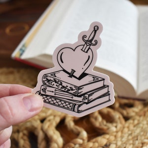 May include: A black and white sticker of a heart pierced by a dagger, sitting on top of a stack of three books. The books have intricate designs on their covers.