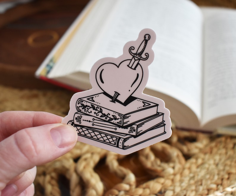 May include: A black and white sticker of a heart pierced by a dagger, sitting on top of a stack of three books. The books have intricate designs on their covers.