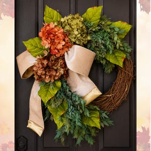 May include: A decorative wreath featuring a circular woven twig base adorned with artificial green leaves, clusters of orange and brown hydrangea flowers, and a large, flowing beige velvet ribbon with gold-toned edges draped across the front.