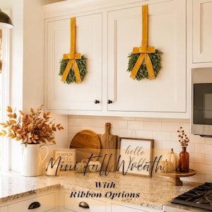 May include: Two small green wreaths hanging on white kitchen cabinets with long mustard yellow velvet ribbons. The text "Mini Fall Wreath With Ribbon Options" is overlaid on the image, along with kitchen decor including pumpkins and signs.