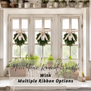 May include: Three small green wreaths hanging on a white window frame, each adorned with a beige and white striped ribbon tied in a bow. The text "Mini Year Round Wreath With Multiple Ribbon Options" is overlaid in black script and print font.