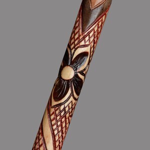 May include: A wooden walking stick with intricate carvings of a flower and geometric patterns. The stick is stained in shades of brown and red.