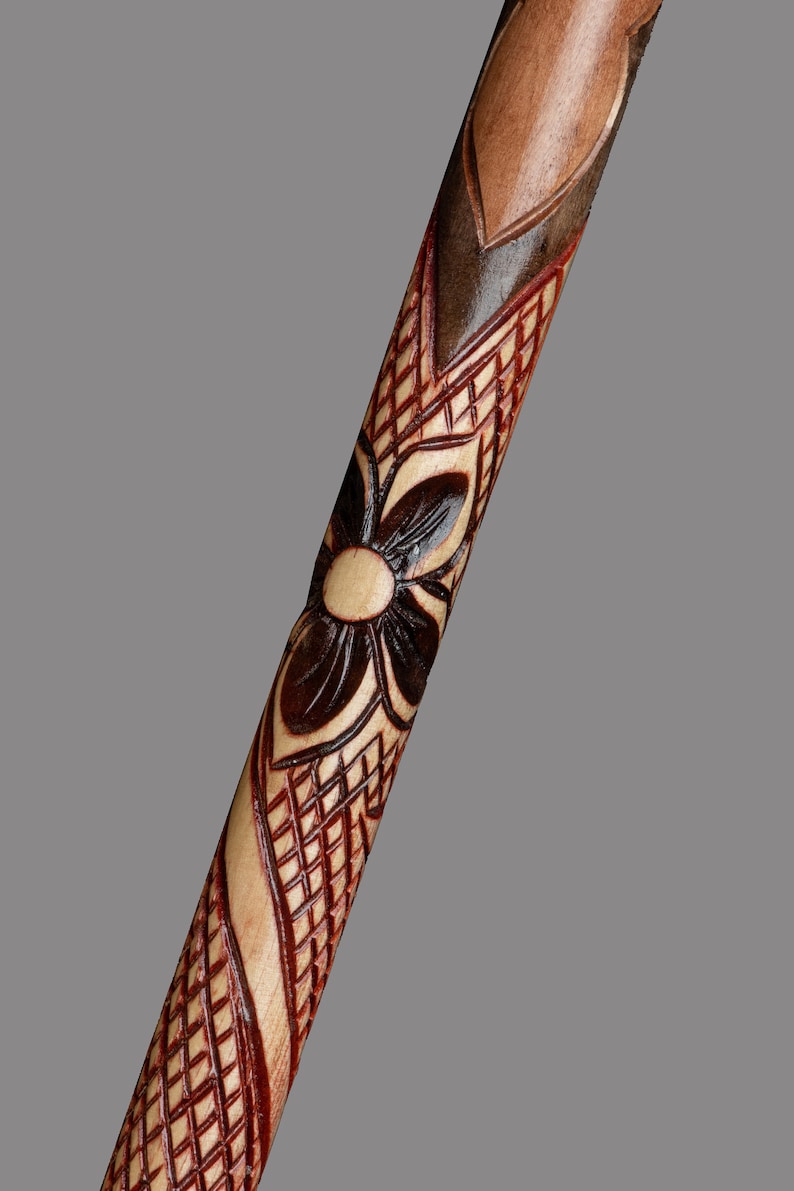 May include: A wooden walking stick with intricate carvings of a flower and geometric patterns. The stick is stained in shades of brown and red.