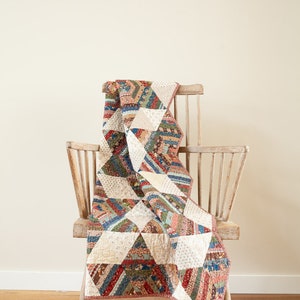 Puede incluir: Una colcha de retazos hecha con recortes de tela de colores está drapeada sobre una silla de madera. La colcha presenta una variedad de patrones y colores, incluyendo rojo, azul, verde y blanco. La silla es de madera clara y tiene un diseño sencillo. La colcha es un gran ejemplo de una "Laundry Basket Quilt", un tipo de colcha hecha con recortes de tela.