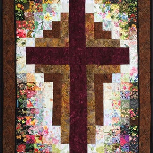 May include: A quilted wall hanging featuring a burgundy cross design at the center. The cross is set against a background of various fabric squares with floral patterns. The quilt has a brown border.
