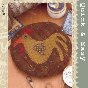 May include: A brown felt chickadee pincushion with a yellow body, red comb, and orange beak. The pincushion is surrounded by green felt grass and brown felt ground. The pincushion is sitting on a wooden table with a spool of thread, a thimble, and a measuring tape.