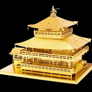 May include: A golden metal model of a Japanese temple with multiple tiers and a detailed roof. The model features intricate architectural details, including balconies and decorative elements. A small figure stands atop the highest point of the temple.