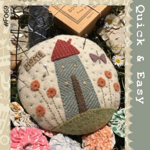 May include: A white felt pincushion shaped like a house with a red roof and a butterfly. The pincushion is surrounded by flowers and has the word "HOME" embroidered on the front.