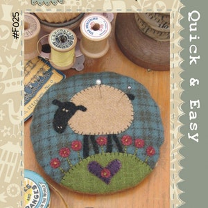 May include: A felt pincushion shaped like a sheep with a heart on a green hill. The sheep is brown with black legs and a black face. The pincushion is surrounded by red felt flowers. The pincushion is labeled "Larry Lamb Pincushion" and "Hatched and Patched #F025".