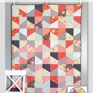 May include: A quilt pattern called "Lofty" featuring a geometric design with a mix of red, pink, gray, black, and white fabrics. The quilt is hanging on a wall with a white chair in the foreground. The logo for "a bright corner" is in the bottom right corner of the image.