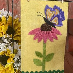 Op de afbeelding: Een gele vlag van vilt voor in de tuin met een roze bloem en een paarse vlinder. De bloem heeft een bruin hart en groene bladeren. De vlag heeft een gegolfde rand met een groene bies.