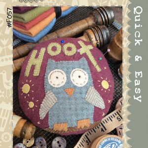 May include: A felt pincushion shaped like an owl with the word "Hoot" stitched around the top. The owl is gray with orange feet and a yellow beak. The pincushion is on a brown surface with a wooden spool and a measuring tape.