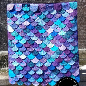 Peut inclure: Une courtepointe bleue et violette avec un motif d'écailles. La courtepointe est étiquetée "Hydra" et "Slice of Pi Quilts".