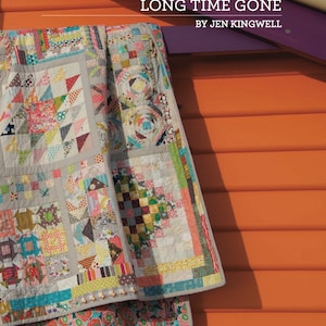 Könnte beinhalten: Ein farbenfroher Patchwork-Decken mit verschiedenen Mustern und Stoffen, der an einer Holzwand mit violettem Rahmen hängt. Die Decke trägt den Titel "Long Time Gone" von Jen Kingwell.