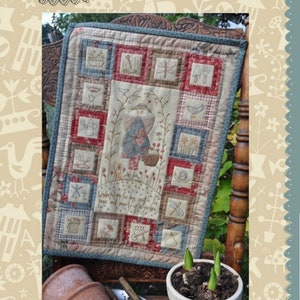 May include: A quilt block with a patchwork design featuring a variety of colors and patterns. The quilt block is surrounded by a blue border and is sitting on a wooden chair. The quilt block has a central image of a woman in a garden with the text "Let your heart find the peace and plenty of the garden". The quilt block is titled "Peaceful Garden" and is part of the "Hatched and Patched" collection.