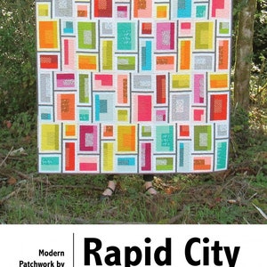 May include: A quilt pattern called "Rapid City" by Elizabeth Hartman. The quilt features a geometric design with colorful squares and rectangles. The quilt is inspired by a mid-century vacation getaway.
