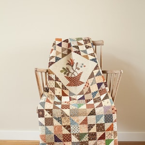 Peut inclure: Une courtepointe en patchwork de couleur marron, beige et bleu avec un motif floral au centre, drapé sur une chaise en bois. La courtepointe est faite de différents tissus et motifs.