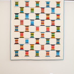 Könnte beinhalten: Ein farbenfroher Quilt mit einem geometrischen Muster aus Garnrollen. Der Quilt hängt an einer weißen Wand mit einem Lichtschalter darunter.