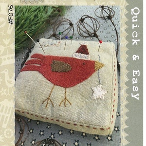 May include: A red felt bird pincushion with a brown felt wing and a white felt star. The bird is wearing a red felt Santa hat. The pincushion is on a beige fabric background with a red stitched border.