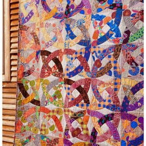 Può includere: Una trapunta colorata con un motivo geometrico di cerchi e archi intrecciati. La trapunta è realizzata con diversi tessuti in tonalità di rosa, viola, blu, arancione, giallo e verde. La trapunta è appesa a una parete di legno con il testo "AARDVARK QUILTS" in basso.
