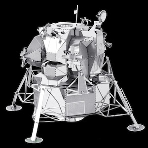 May include: A detailed, silver-colored metal model of the Apollo Lunar Module, featuring intricate paneling, antennas, and landing gear. The model is set against a black background, showcasing its complex design and construction. This item is a collectible or a display piece.
