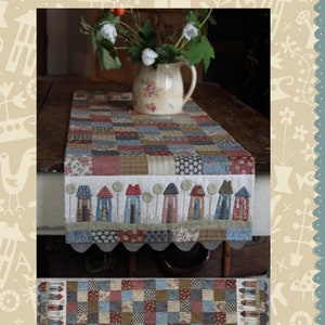 May include: A quilted table runner with a patchwork design featuring a row of houses and trees. The table runner is gray, blue, red, and brown. The text "Tiny Town Tablerunner" is at the bottom of the image.