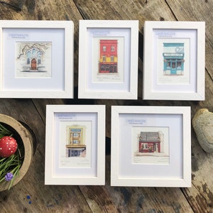 Könnte beinhalten: Fünf weiße gerahmte Drucke von Aquarell-Illustrationen von Gebäuden in einer Stadt. Jeder Druck hat einen Titel und den Namen des Künstlers, "Craftworks Art".
