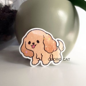 Puede incluir: Una pegatina de un perro de dibujos animados con un pelaje marrón claro y una lengua rosa que sobresale. El perro tiene un acabado brillante e iridiscente.