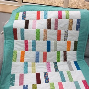 May include: A white quilt with a teal border. The quilt is made of various colorful fabric strips sewn together in a patchwork pattern.