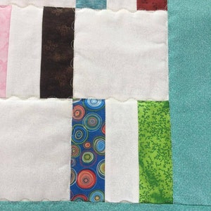 May include: A close-up of a quilt with a variety of colorful fabric squares. The squares are sewn together in a patchwork pattern. The quilt is made with a variety of fabrics, including floral prints, geometric patterns, and solid colors. The quilt is edged with a teal fabric.