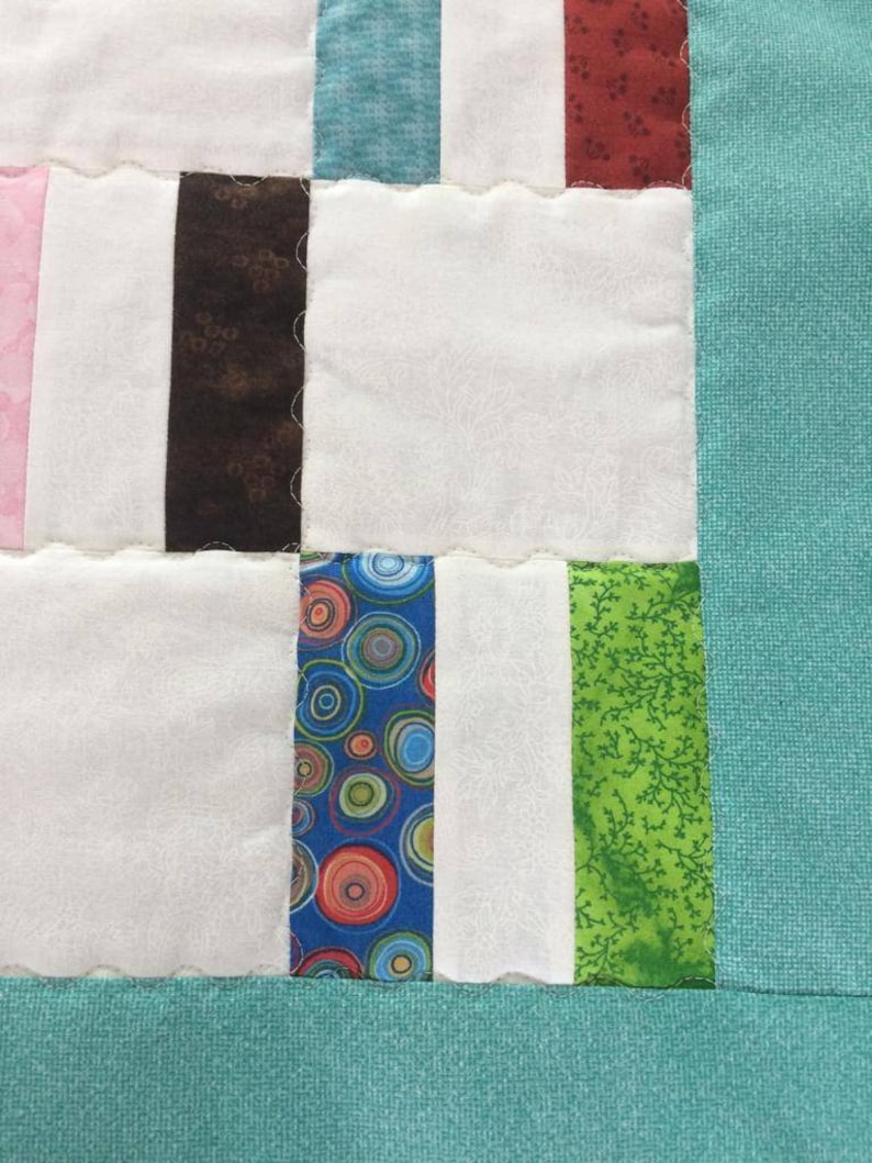 May include: A close-up of a quilt with a variety of colorful fabric squares. The squares are sewn together in a patchwork pattern. The quilt is made with a variety of fabrics, including floral prints, geometric patterns, and solid colors. The quilt is edged with a teal fabric.
