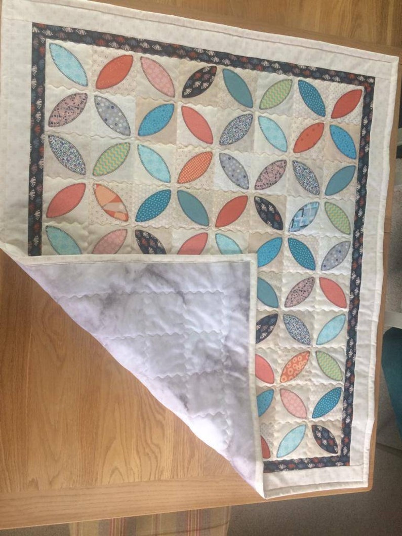 Pode incluir: Um edredom branco com um padr&atilde;o geom&eacute;trico de ovais sobrepostos em azul-petr&oacute;leo, laranja e rosa. O edredom est&aacute; dobrado ao meio, revelando um forro branco.