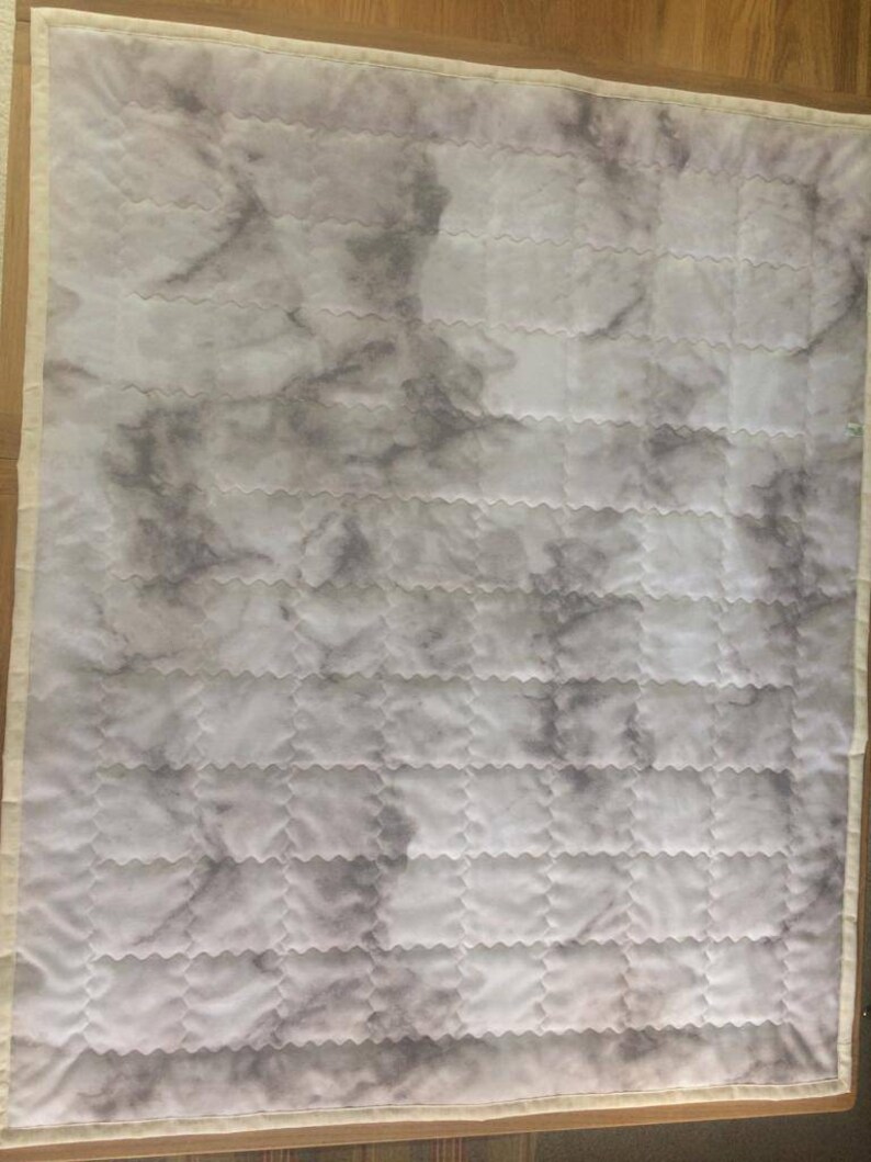 Pode incluir: Uma colcha branca com um padr&atilde;o de m&aacute;rmore cinza. A colcha &eacute; acolchoada em um padr&atilde;o de diamante com uma borda festoneada.