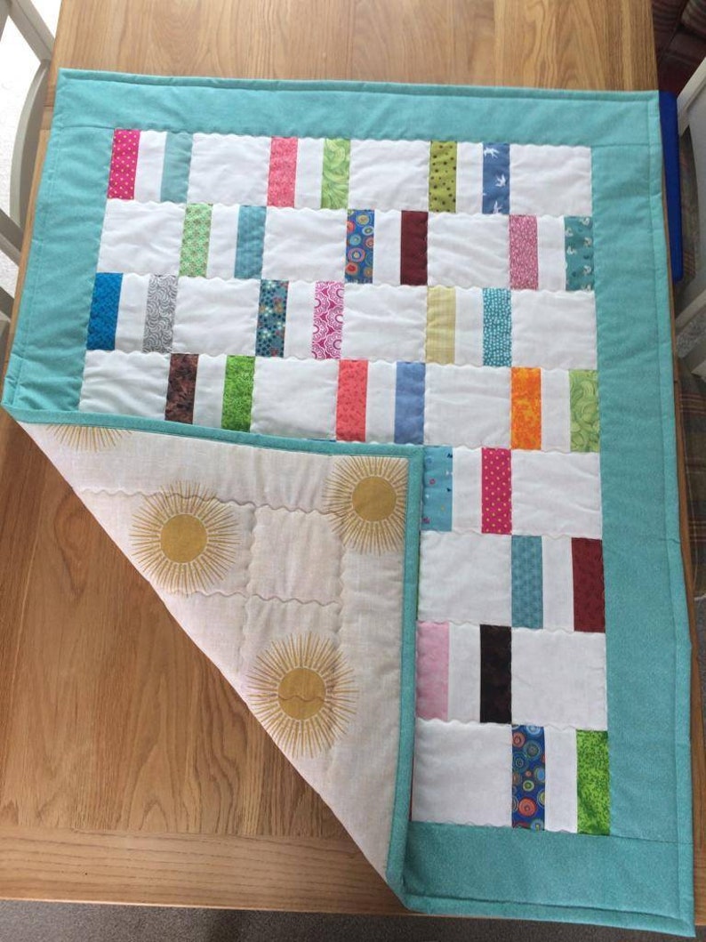 May include: A turquoise blue quilted throw with a white background and colorful fabric squares. The back of the throw is white with a sun design.