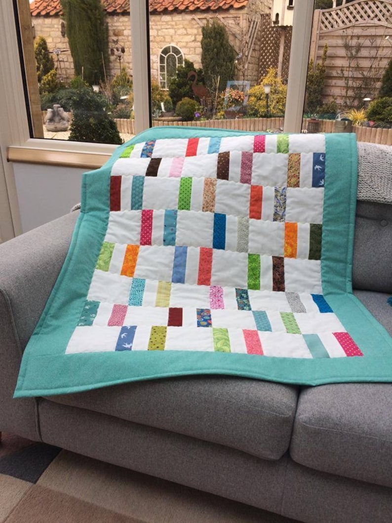 May include: A teal blue quilt with a white background and colorful patchwork squares. The quilt is folded and sitting on a gray couch.