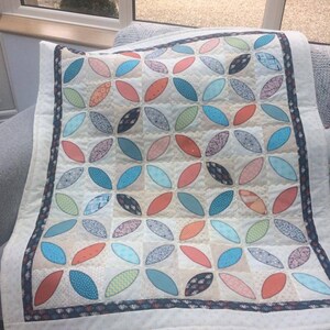 Pode incluir: Um edredom branco com um padr&atilde;o geom&eacute;trico de c&iacute;rculos sobrepostos turquesa, laranja e verde. O edredom tem uma borda preta e branca.