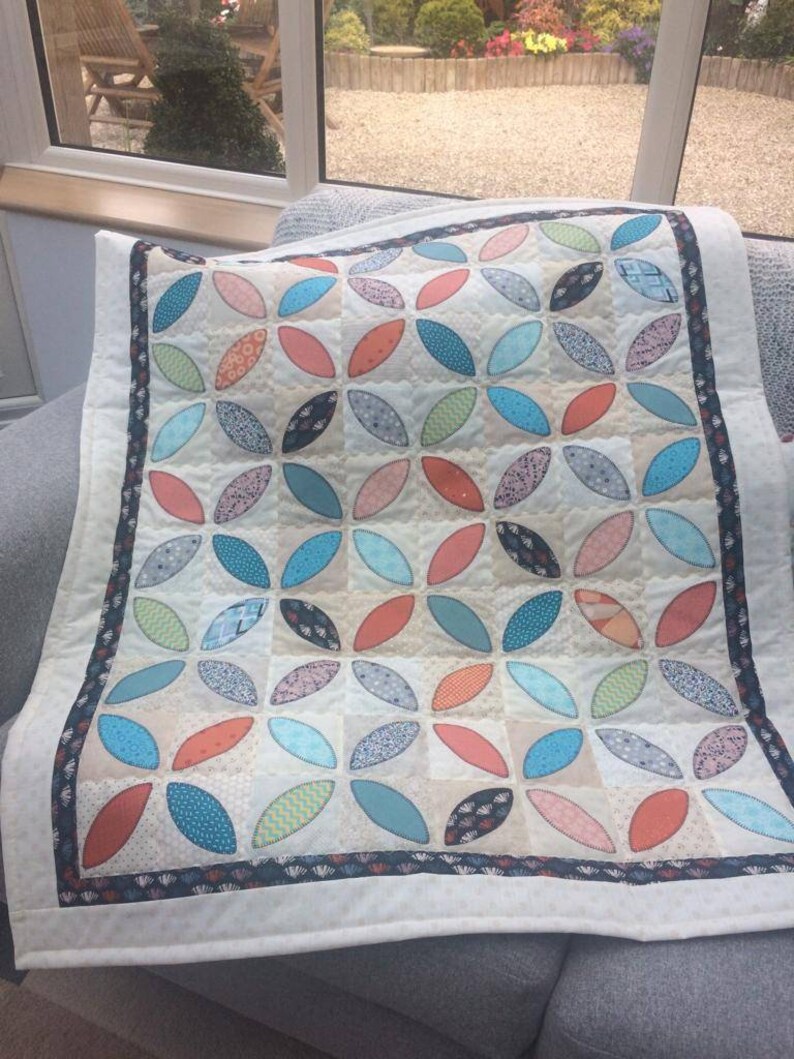 Pode incluir: Um edredom branco com um padr&atilde;o geom&eacute;trico de c&iacute;rculos sobrepostos turquesa, laranja e verde. O edredom tem uma borda preta e branca.