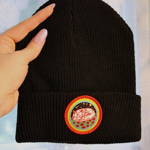May include: Black beanie with a colorful embroidered patch of a cat sleeping on a bed of flowers. The patch has a rainbow border and stars.