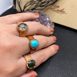 May include: Three gold rings with different colored stones. The rings are on a hand, and the stones are green, blue, and a mottled green and white.
