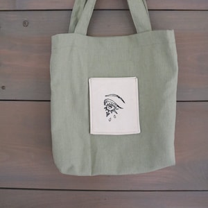 Puede incluir: Una bolsa de lona verde con un parche blanco que presenta un diseño de ojo en blanco y negro.