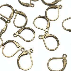 May include: A group of 15 gold-toned earring clasps, also known as earring hooks, are scattered on a white surface. The clasps are made of metal and have a teardrop shape.