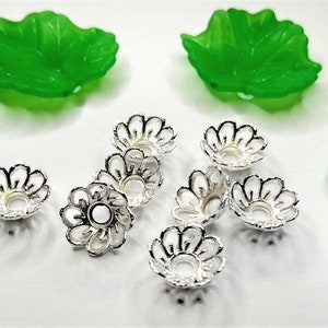 May include: An assortment of jewelry-making components, including bright green leaf-shaped beads and silver-colored flower-shaped bead caps. The bead caps have a detailed, openwork design. The components are arranged on a white surface.