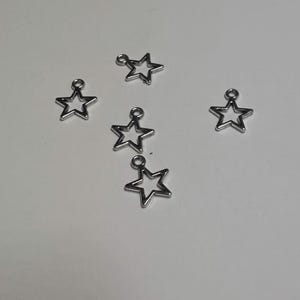 May include: Five small, silver-colored metal star charms, each featuring an open-center design and a circular jump ring at the top point for attachment. The charms are arranged in a scattered pattern, showcasing their metallic finish and star shape.