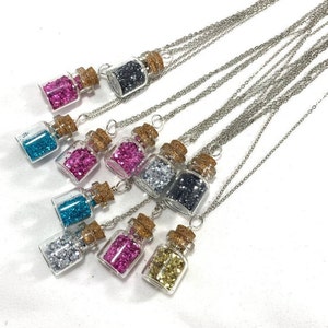 Puede incluir: Un conjunto de diez collares de cadena plateada con pequeñas botellas de vidrio llenas de purpurina de diferentes colores. Cada botella tiene un tapón de corcho.