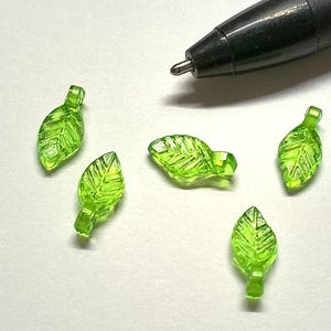 May include: Five green glass leaf charms with a textured surface. The charms have a hole at the top for attaching to jewelry or other crafts.