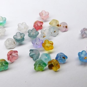 Puede incluir: Un conjunto de pequeñas cuentas con forma de flor en varios colores, incluyendo verde, rosa, azul y amarillo. Las cuentas parecen ser de vidrio o un material translúcido similar, algunas con detalles dorados. Están dispersas sobre una superficie blanca.