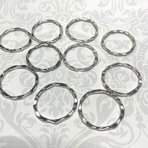 May include: A collection of ten silver-colored, wavy-edged metal rings. The rings are circular with an irregular, textured surface. They are scattered on a patterned white surface, suggesting a craft or jewelry-making theme.